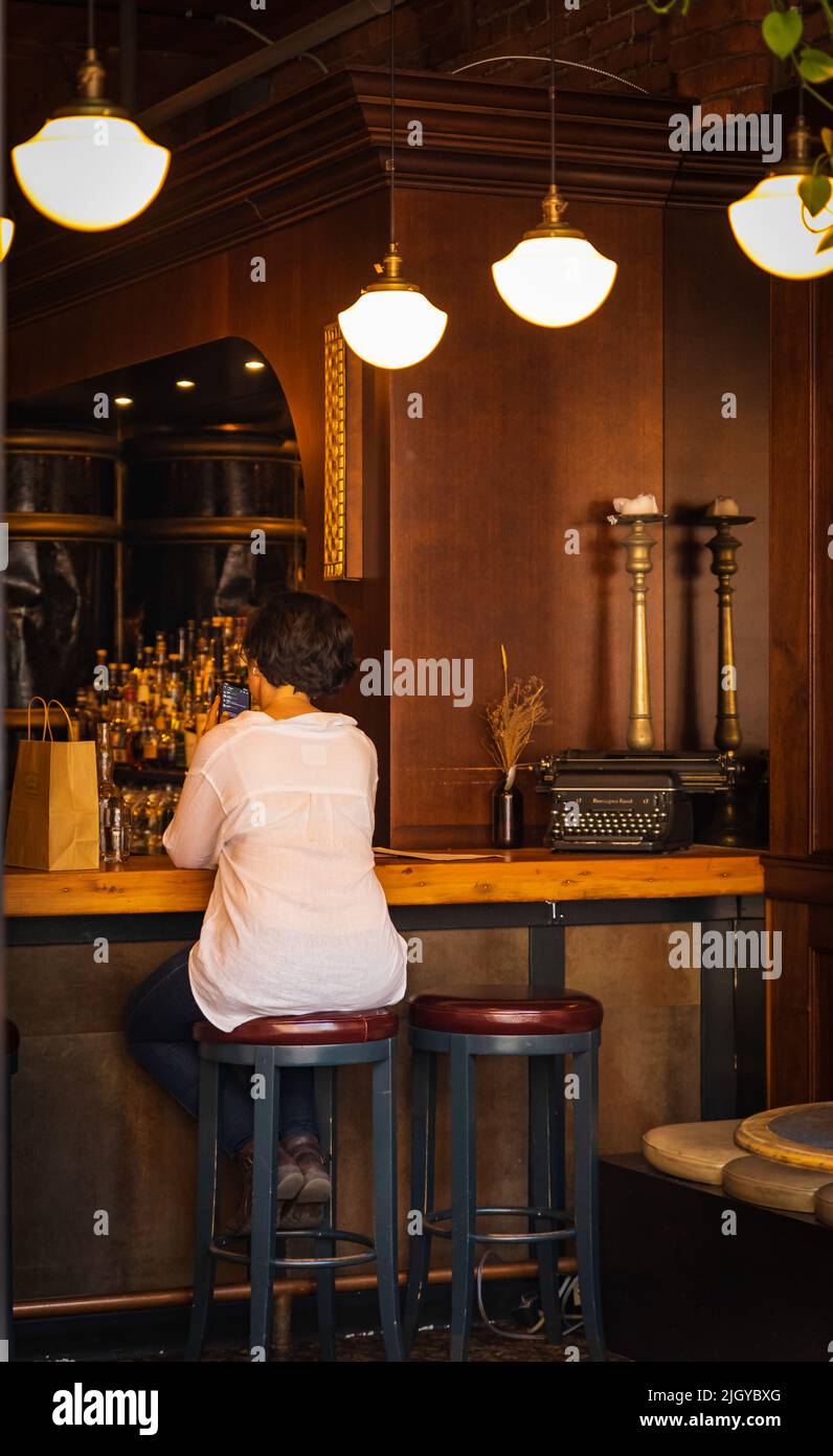 Bella donna sta ordinando un drink al bar, vista dal retro. Bere e aspettare. Donna ricci dai capelli lunghi che beve vino e aspetta il suo ma Foto Stock