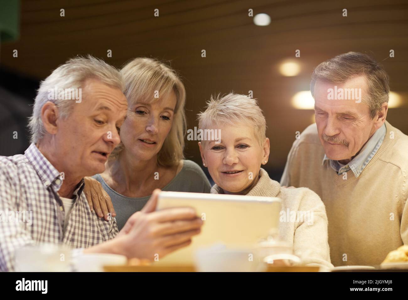 Gruppo di amici in pensione positivi in abbigliamento casual seduti a tavola e utilizzando un tablet digitale mentre si guardano i video online insieme Foto Stock