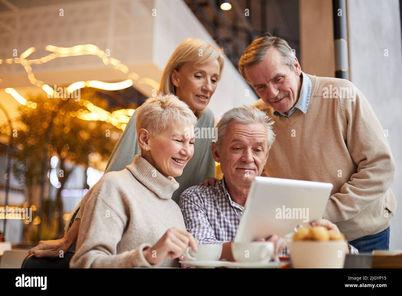 Gruppo di amici senior positivi in abbigliamento casual utilizzando tablet mentre navigano siti web in un bar con wifi Foto Stock