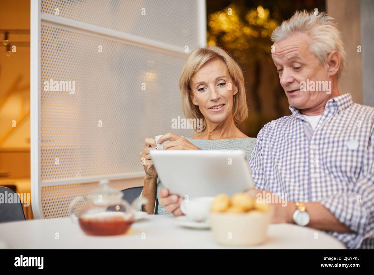 Positivo bella coppia senior seduta al tavolo con tè e dolci e l'uso di dispositivi moderni per guardare il video Foto Stock