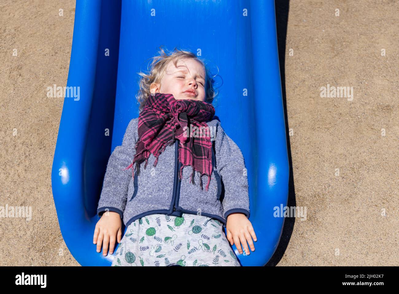 Divertente ritratto dall'alto verso il basso di ragazzo caucasico di due anni, con capelli biondi ondulati che fingono di dormire con gli occhi chiusi giacenti sul fondo dello scivolo del parco giochi. Foto Stock
