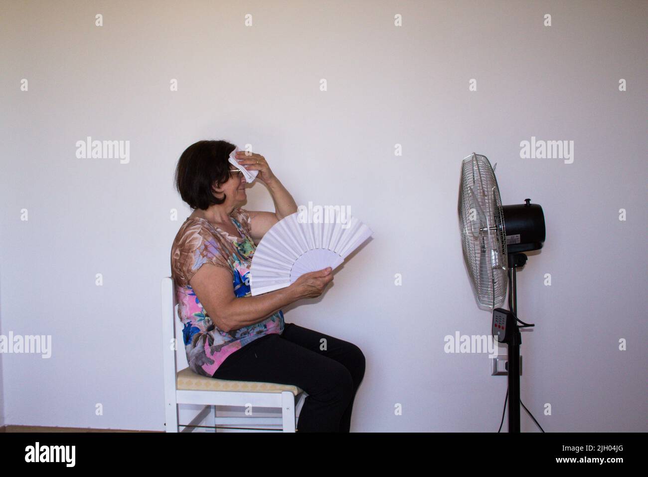 Immagine di una donna anziana seduta su una sedia che puliva il sudore con un fazzoletto e cerca rinfresco con un ventilatore e un richiamo. Riferimento al suffe Foto Stock