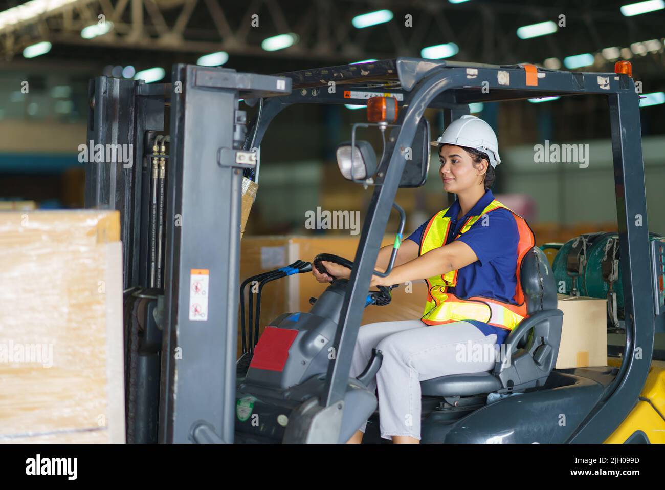 Asian bella donna conducente guida carrello elevatore auto in industria con sorriso, abilità della ragazza e diversità di carriera indossare casco in maglia riflettente Foto Stock