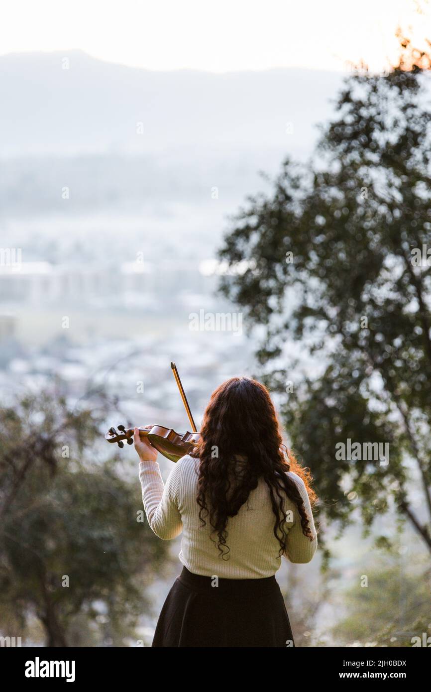 Donna bruna vista dalla parte posteriore giocando il violino fuori nella montagna, e il paesaggio urbano. Verticale Foto Stock