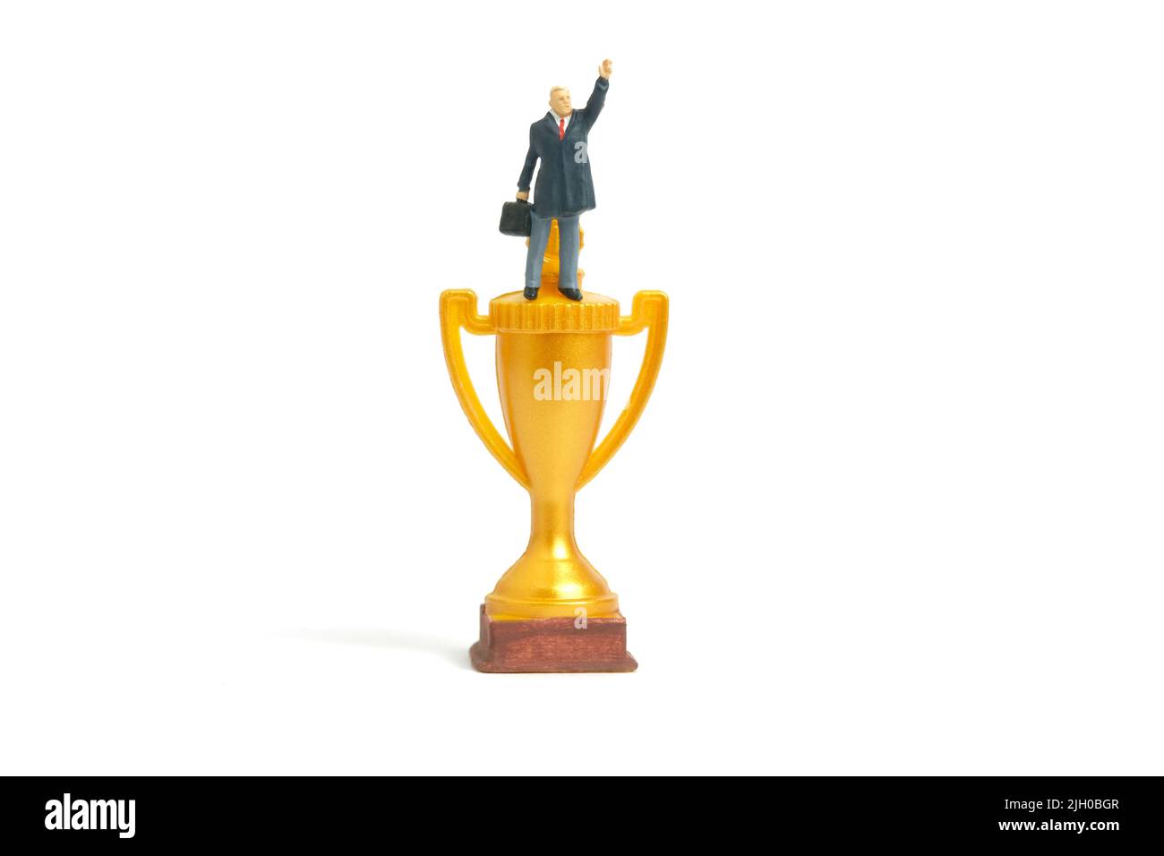 Fotografia di personaggi giocattolo in miniatura. Un uomo d'affari che si trova al di sopra del trofeo d'oro determina di vincere o diventare un vincitore mentre alza la mano. Isolato o Foto Stock