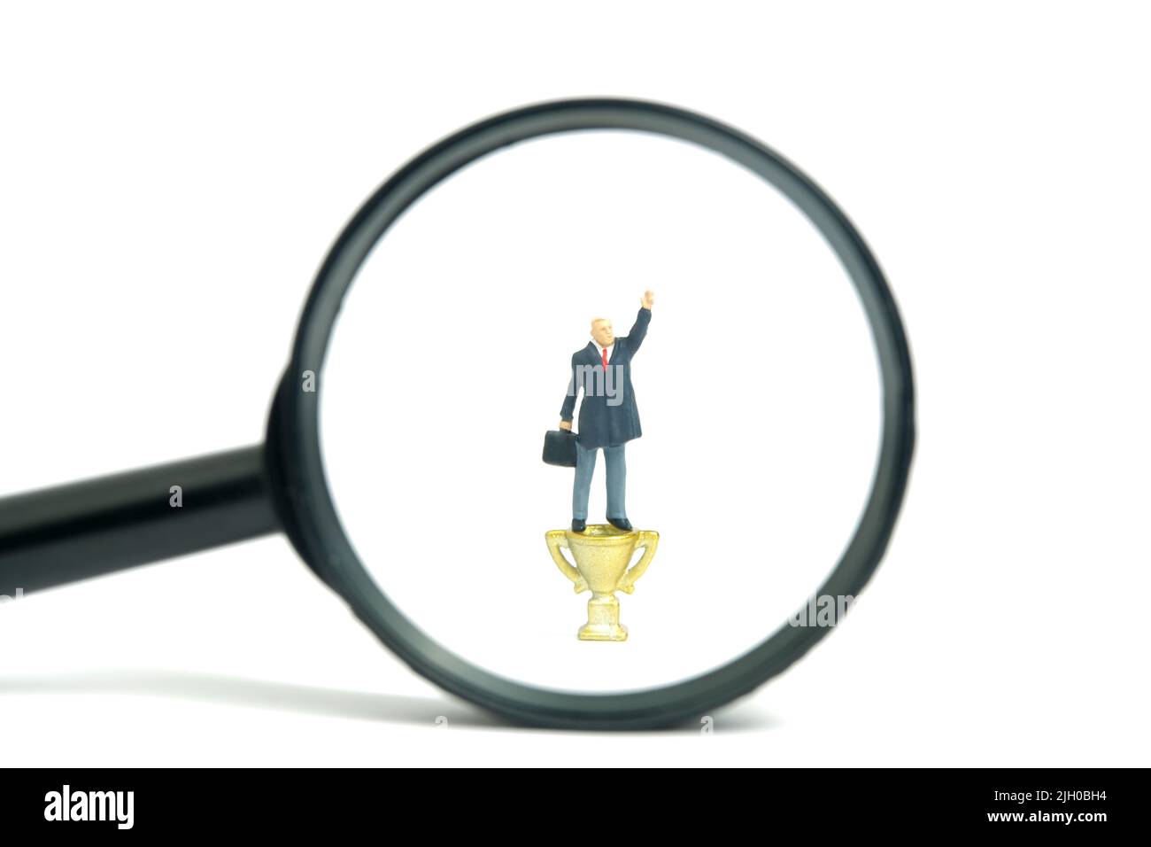Fotografia di personaggi giocattolo in miniatura. Un uomo d'affari in piedi sopra il trofeo dorato dietro la lente d'ingrandimento determina diventare un vincitore. Isolato su w Foto Stock