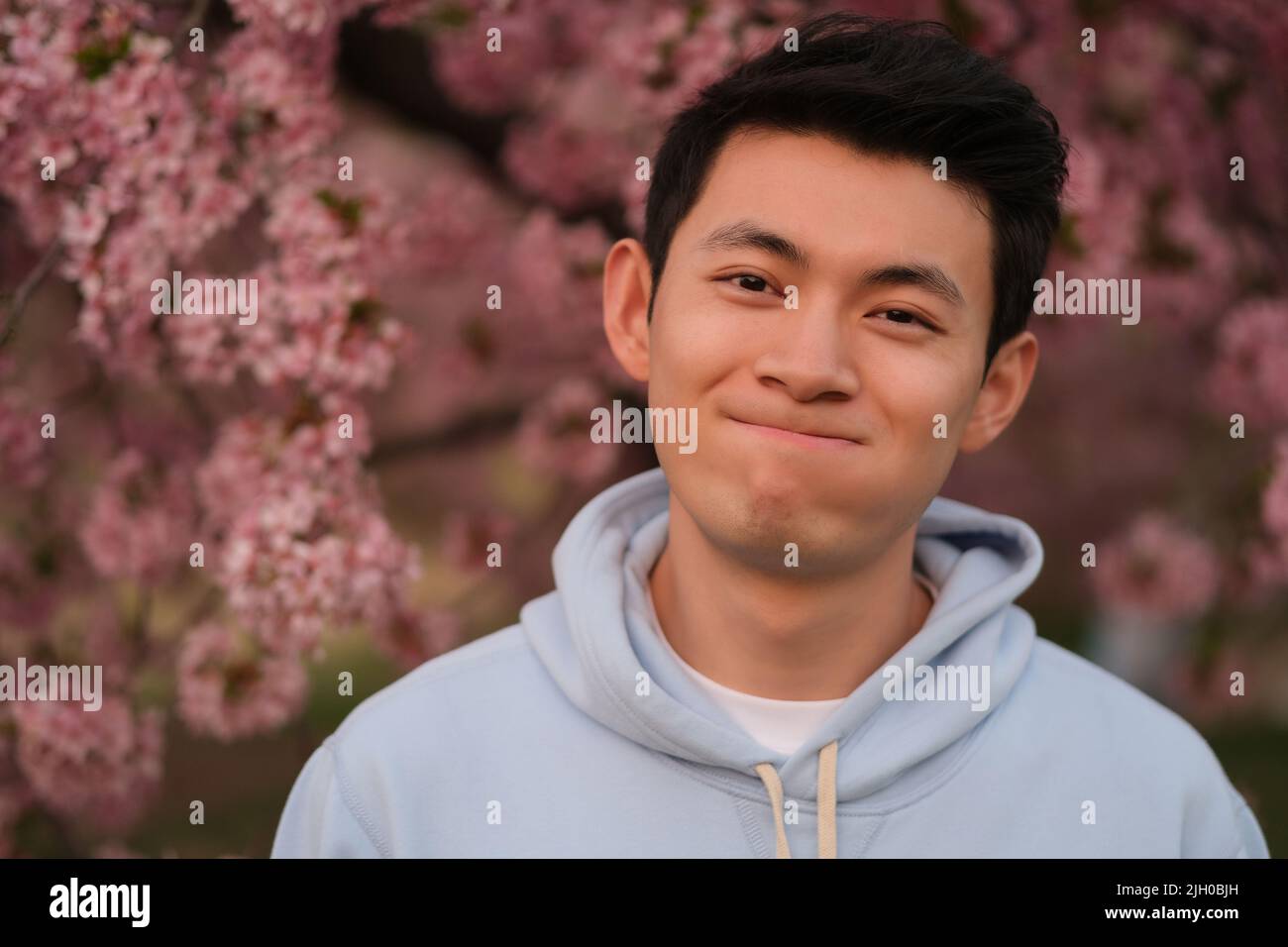 Grazioso giovane asiatico che fa il viso, guardando la macchina fotografica. Sfocatura rosa sakura fiori sfondo Foto Stock