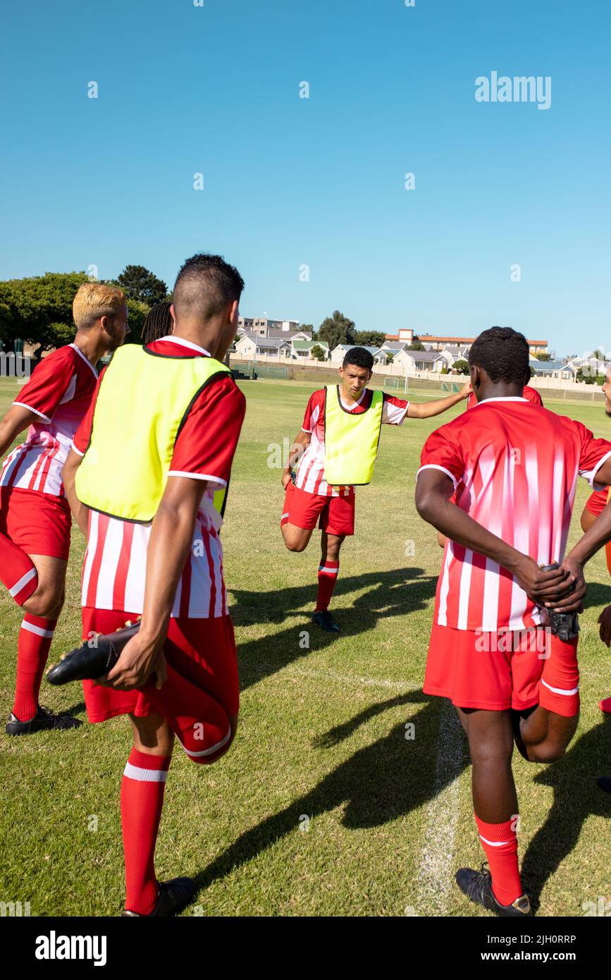 Atleti maschi multirazziali che indossano uniformi rosse che allungano le gambe al parco giochi contro il cielo limpido Foto Stock