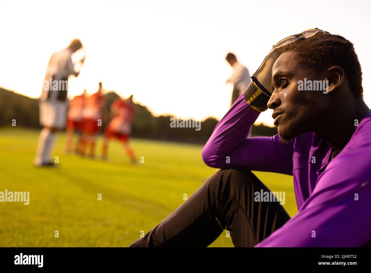 African american ha sottolineato il portiere maschile con la testa in mano seduta sul campo contro il cielo limpido Foto Stock