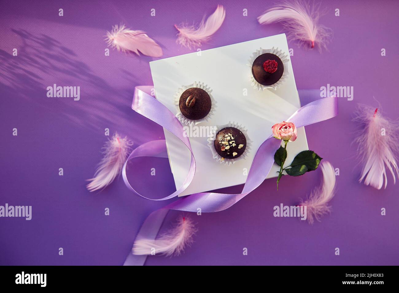 Vista dall'alto delle caramelle al cioccolato natalizie con ombre. Delicate piume rosa e nastro viola. Sfondo con motivo di colore viola. Foto Stock