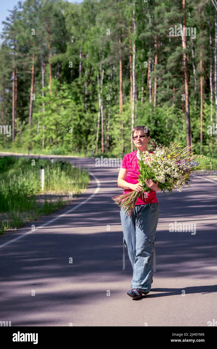 ritratto a tutta lunghezza di una donna sorridente felice con un enorme bouquet di fiori selvatici che camminano lungo una strada forestale Foto Stock