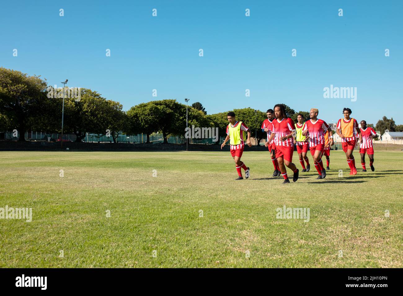 Squadra di calcio maschile multirazziale che indossa uniformi rosse che corrono su terra erbosa contro il cielo limpido Foto Stock