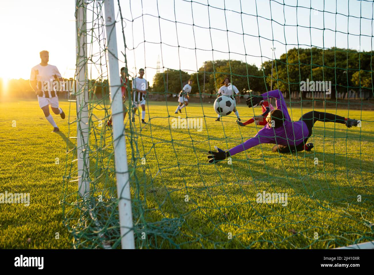 Portiere multirazziale che difende la palla di obiettivo avversario durante la partita contro il cielo chiaro al tramonto Foto Stock