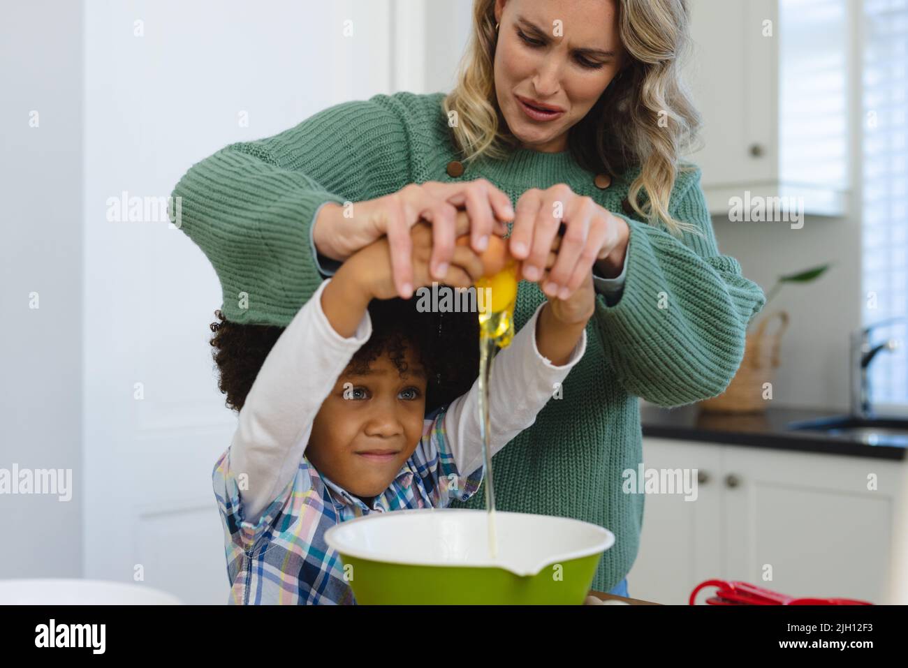 Madre multirazziale che fa il viso mentre aiuta il figlio con capelli afro in rottura uovo in ciotola in cucina Foto Stock