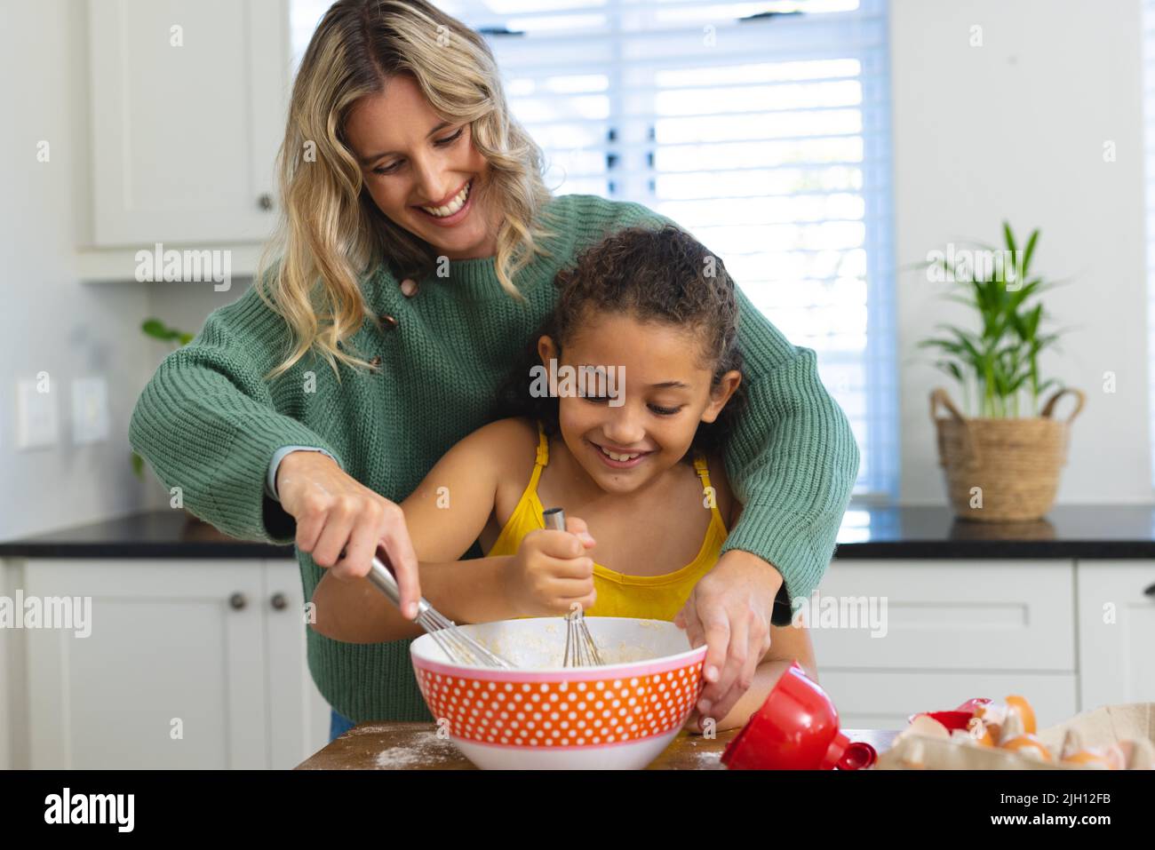 Allegro madre multirazziale insegnamento figlia cute per mescolare pastella con filo frusta in ciotola in cucina Foto Stock