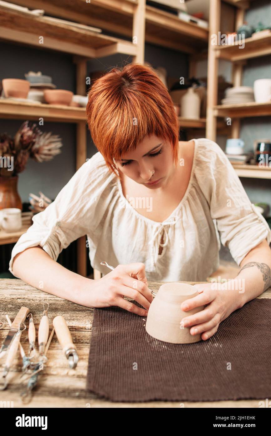 Giovane artigiano moderno che decora la ceramica. Bella donna con tatuaggio e body piercing carving vaso in argilla in officina, hobby di generazione moderna Foto Stock