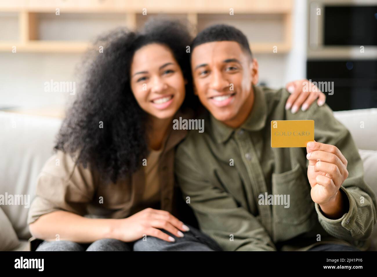 Felice giovane coppia sorridente con piacere, mentre si siede sul divano con carta di credito e guardando la macchina fotografica. Felice uomo biraciale mostrando carta di credito di plastica. Concetto di shopping online Foto Stock