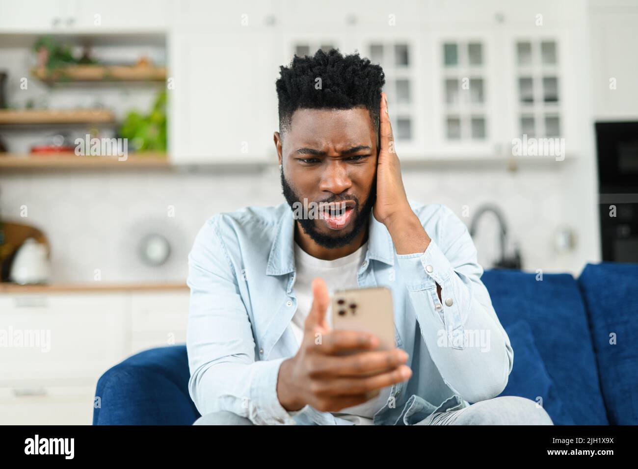 Arrabbiato ha stressato uomo multirazziale che ha sgradevole conversazione di videochiamata, parlare su smartphone online e discutere, mentre si siede sul divano e guardando lo schermo del telefono con espressione del volto frustrata Foto Stock