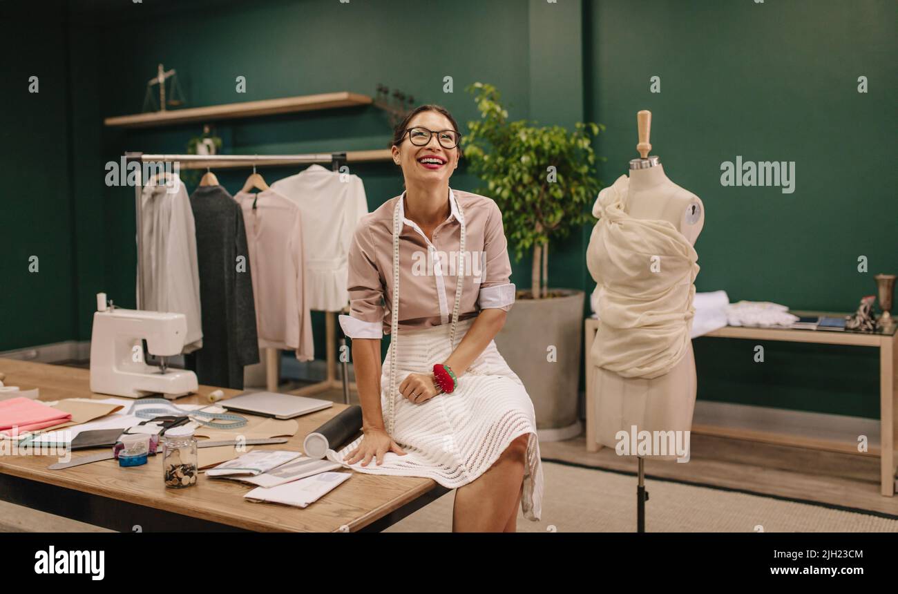Designer asiatico sorridente e seduto sul tavolo nel suo studio. Designer femminile al suo posto di lavoro. Foto Stock
