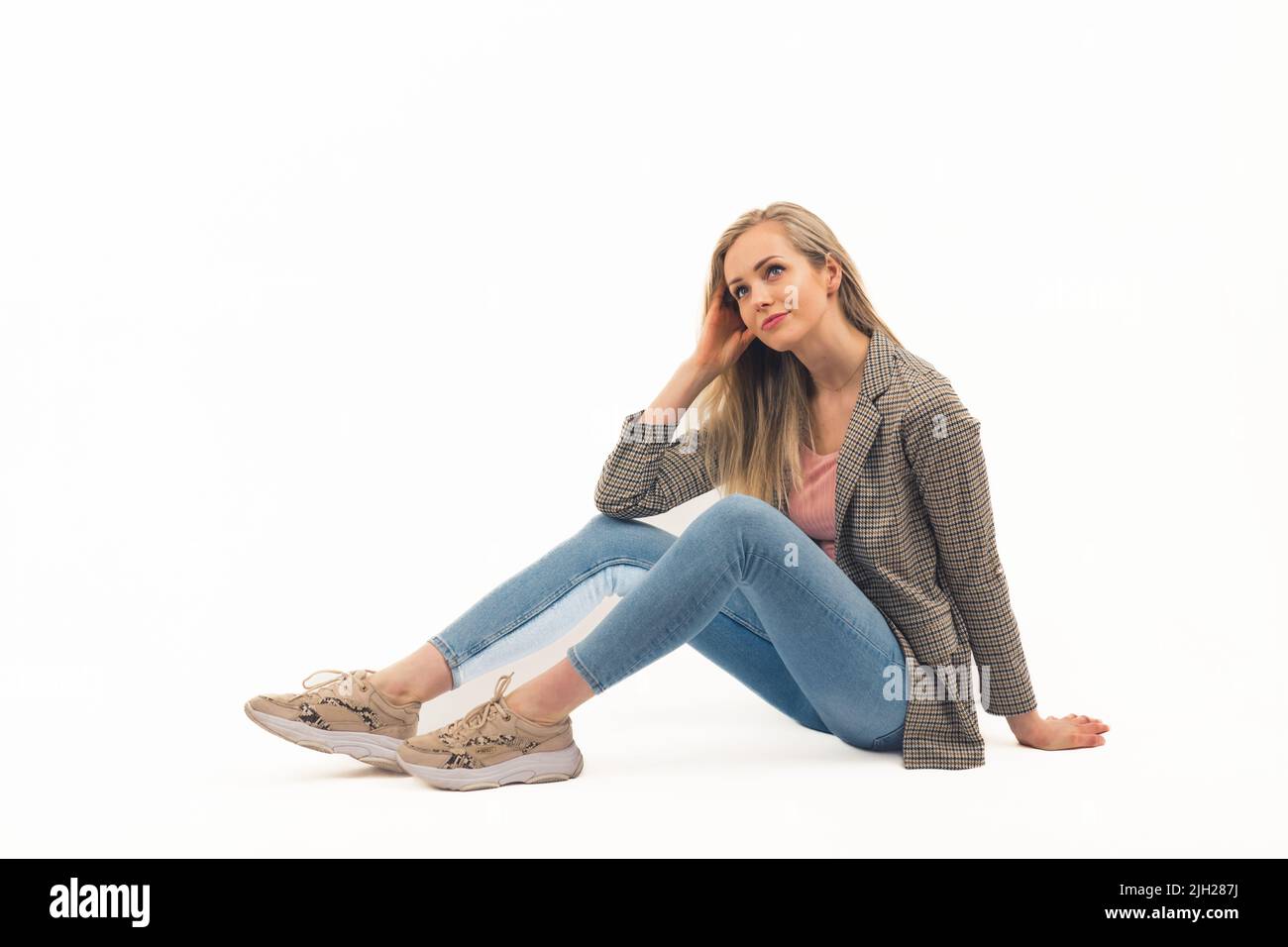 Giovane donna bionda adulta vestita in jeans e blazer controllato seduto a terra su sfondo bianco e guardando in su con un sorriso morbido. Foto studio dello spazio di copia isolato. Foto di alta qualità Foto Stock