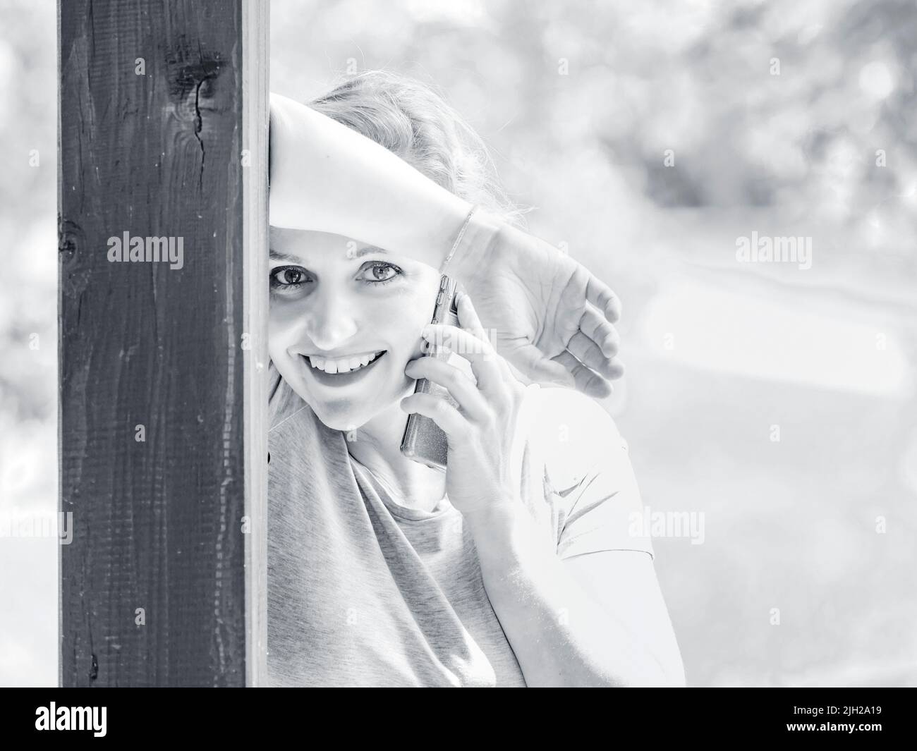 Occhi capelli biondi donna 30s sta parlando su smartphone inclinato su colonna di legno montante felice sorridente guardando la fotocamera high-hey highkey monocromatico Foto Stock