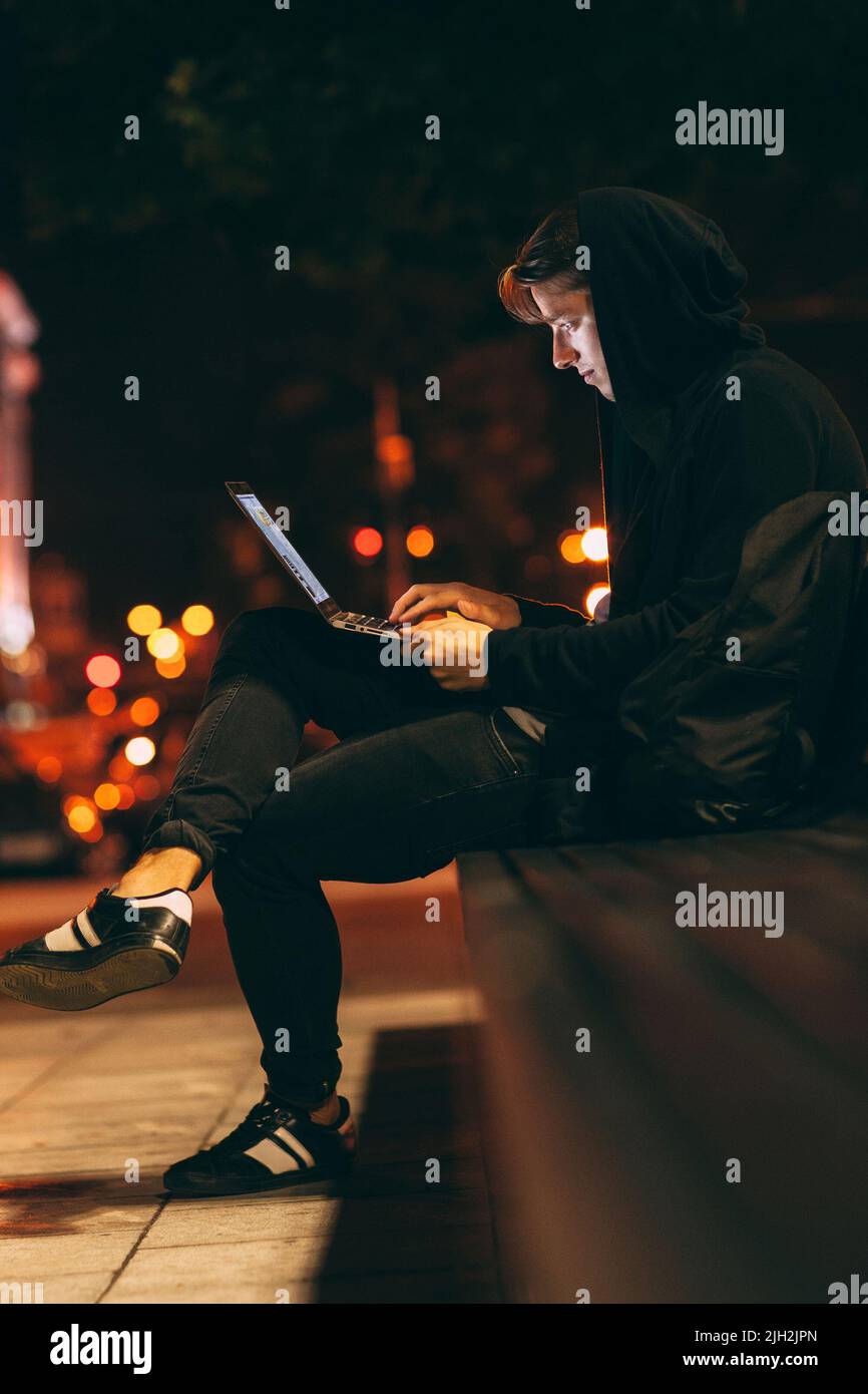 Uomo con cappuccio seduto su una panca con computer portatile Foto Stock