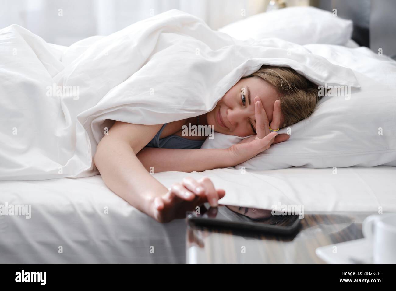 Donna addormentata coperta di coperta di coperta la mattina a letto che spegne la sveglia su cellulare. È difficile svegliarsi presto dal letto Foto Stock