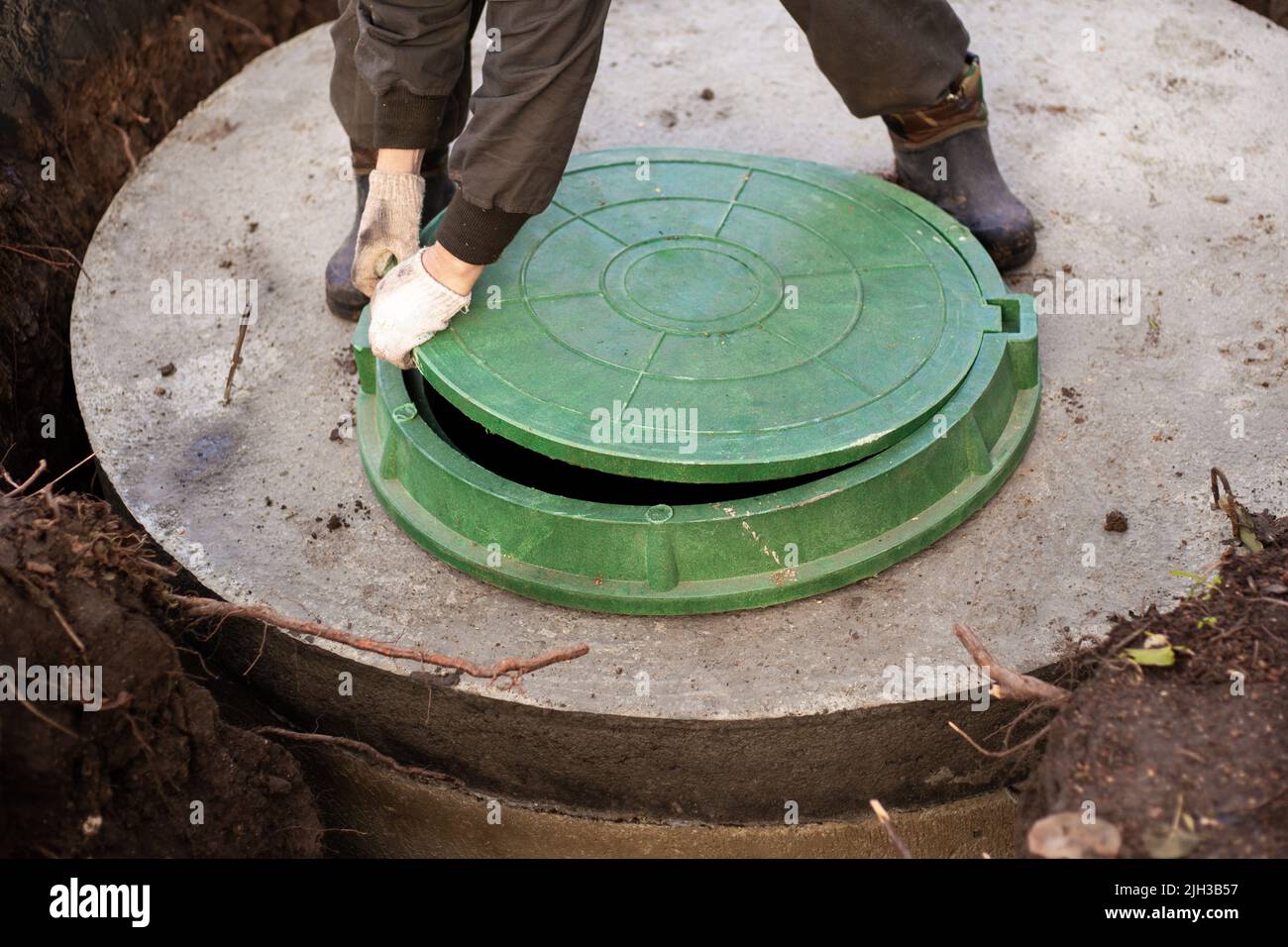 Un lavoratore installa un tombino fognario su un serbatoio settico fatto di anelli di cemento. Costruzione di reti fognarie per case di campagna. Foto Stock