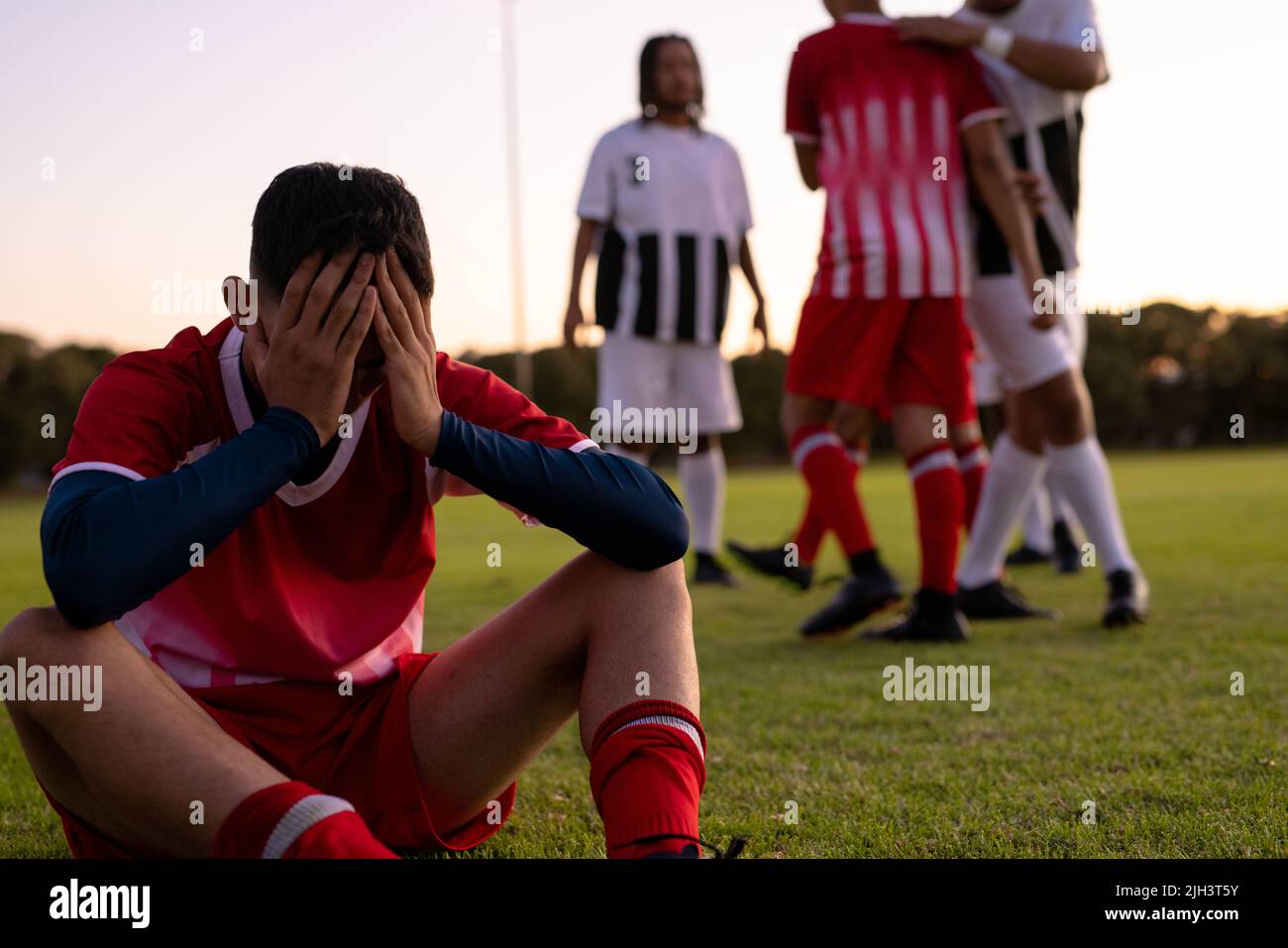 Giocatore di calcio maschile multirazziale triste con testa in mano seduta su terra erbosa contro cielo chiaro Foto Stock