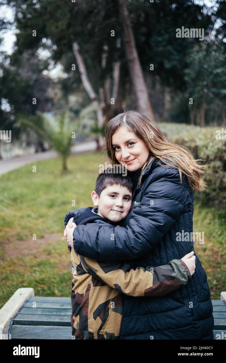 Giovane madre bionda che abbraccia il suo giovane figlio in una strada del parco. Famiglia con un solo genitore Foto Stock