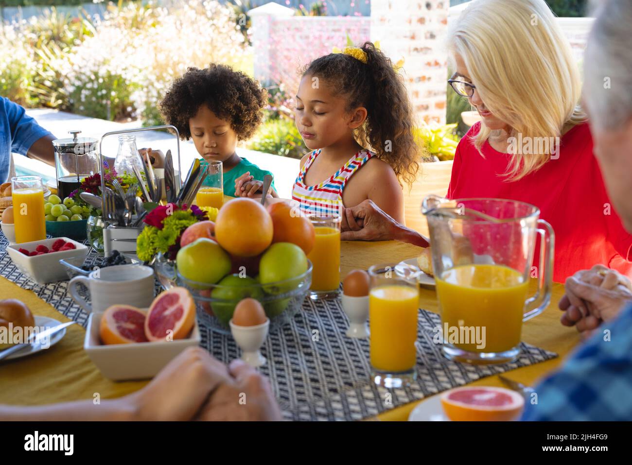 Nonna multirazziale e nipoti che tengono le mani e pregano prima della colazione al tavolo da pranzo Foto Stock