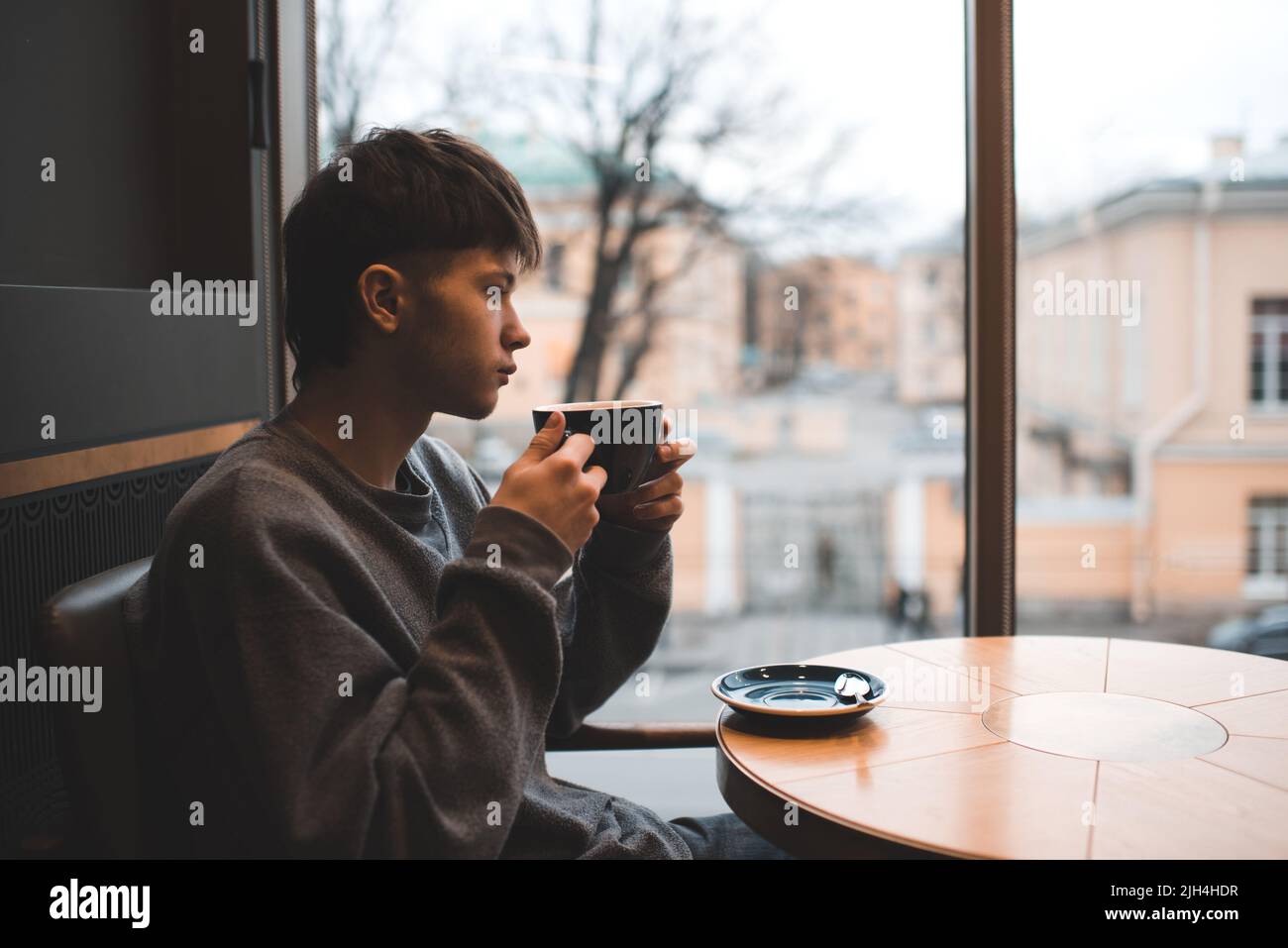 Giovane bel ragazzo di 18-19 anni bevendo caffè seduto in un caffè da solo con un triste umore abbandonato. Stile di vita della generazione Z e dei Millennial. Foto Stock