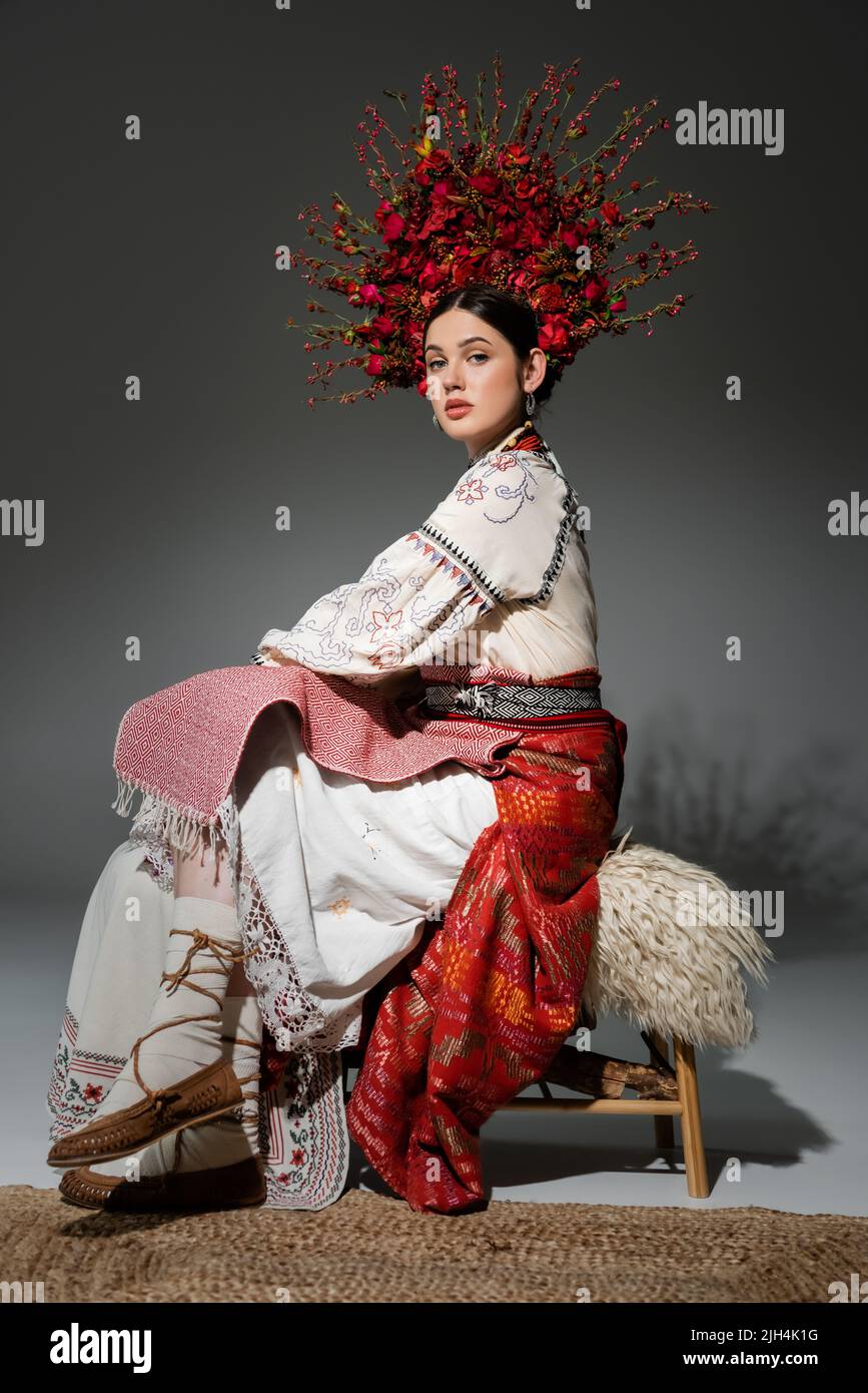 tutta la lunghezza della donna ucraina graziosa in vestiti tradizionali e la corona rossa con i fiori e bacche che siedono sul grigio Foto Stock