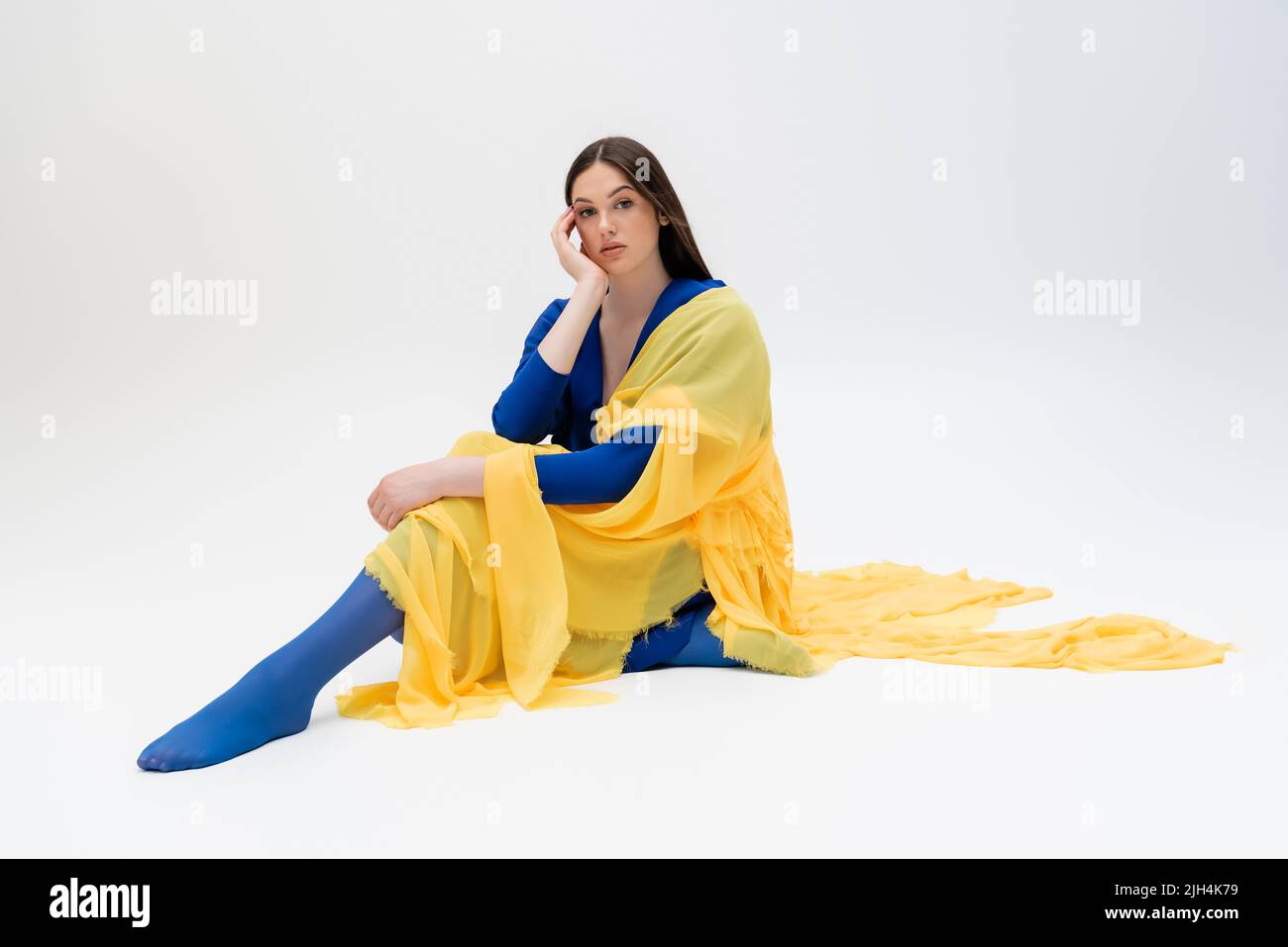 giovane donna ucraina sognante in abito blu e giallo con collant seduti in posa sul grigio Foto Stock