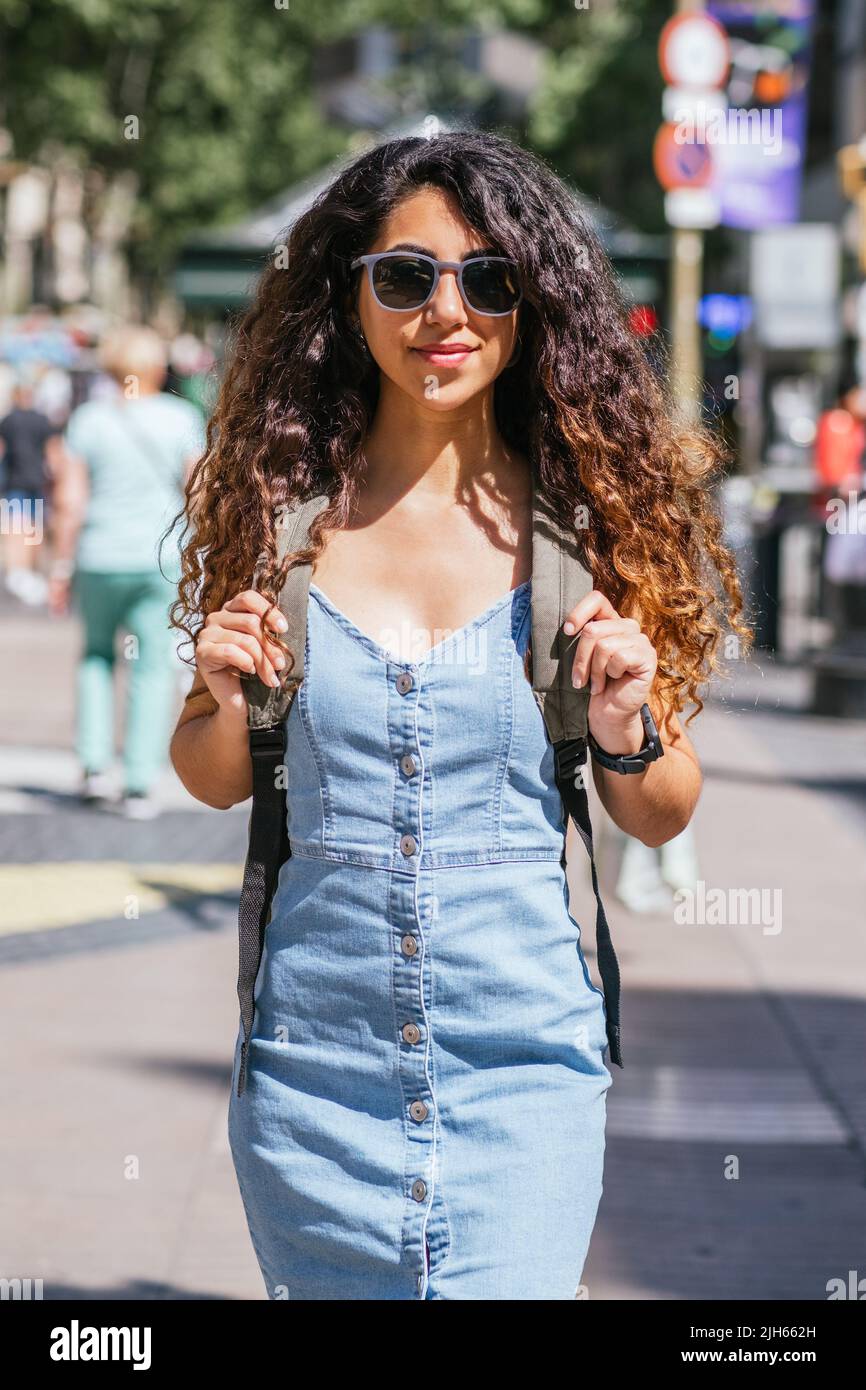 attraente turista dai capelli scuri che cammina per la strada con uno zaino. Indossa un abito in denim e occhiali da sole Foto Stock