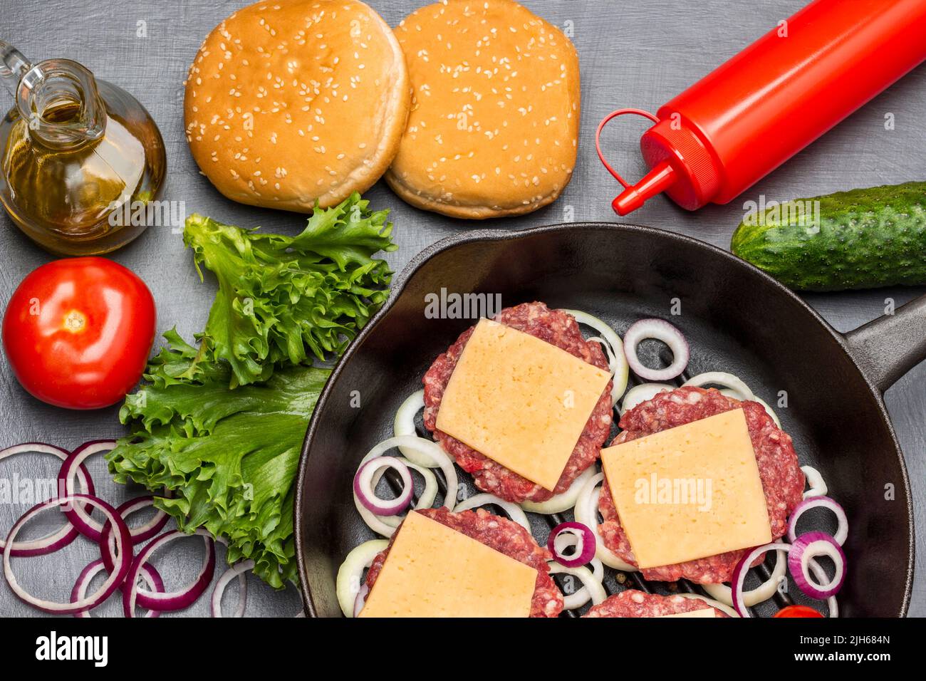 Panini con insalata verde di sesamo, pomodoro, cetriolo e olio in bottiglia di vetro. Hamburger di manzo crudo con formaggio e cipolle tritate in padella. Disposizione piatta. Scuro Foto Stock