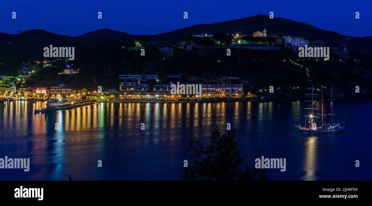 Vista panoramica di Porto Azzurro con passeggiata del porto illuminata di sera durante l'ora blu, in alto a destra sullo sfondo Casa di Reclusione Foto Stock