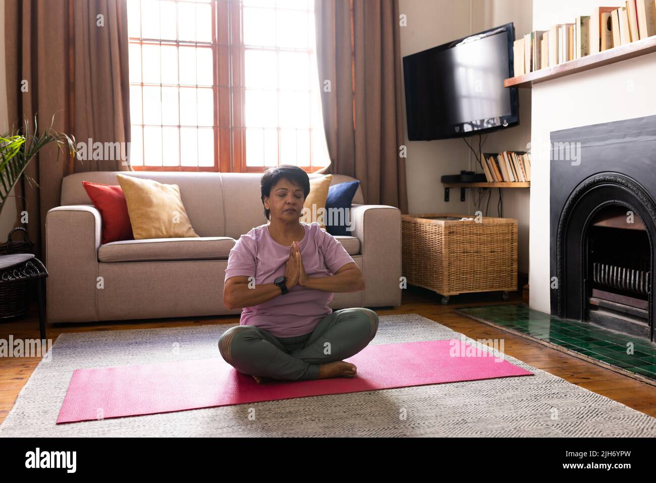 Donna matura biraciale con capelli corti che chiudono gli occhi e meditando in posizione di preghiera sul tappeto a casa Foto Stock