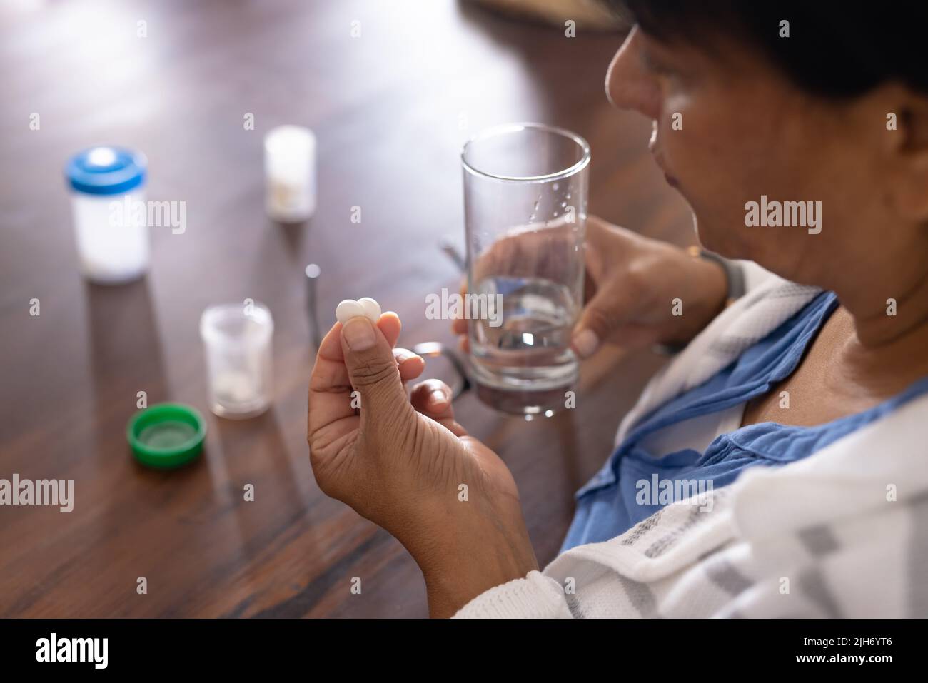 Vista ad alto angolo della donna matura biraciale che tiene medicinali e acqua potabile a tavola a casa Foto Stock