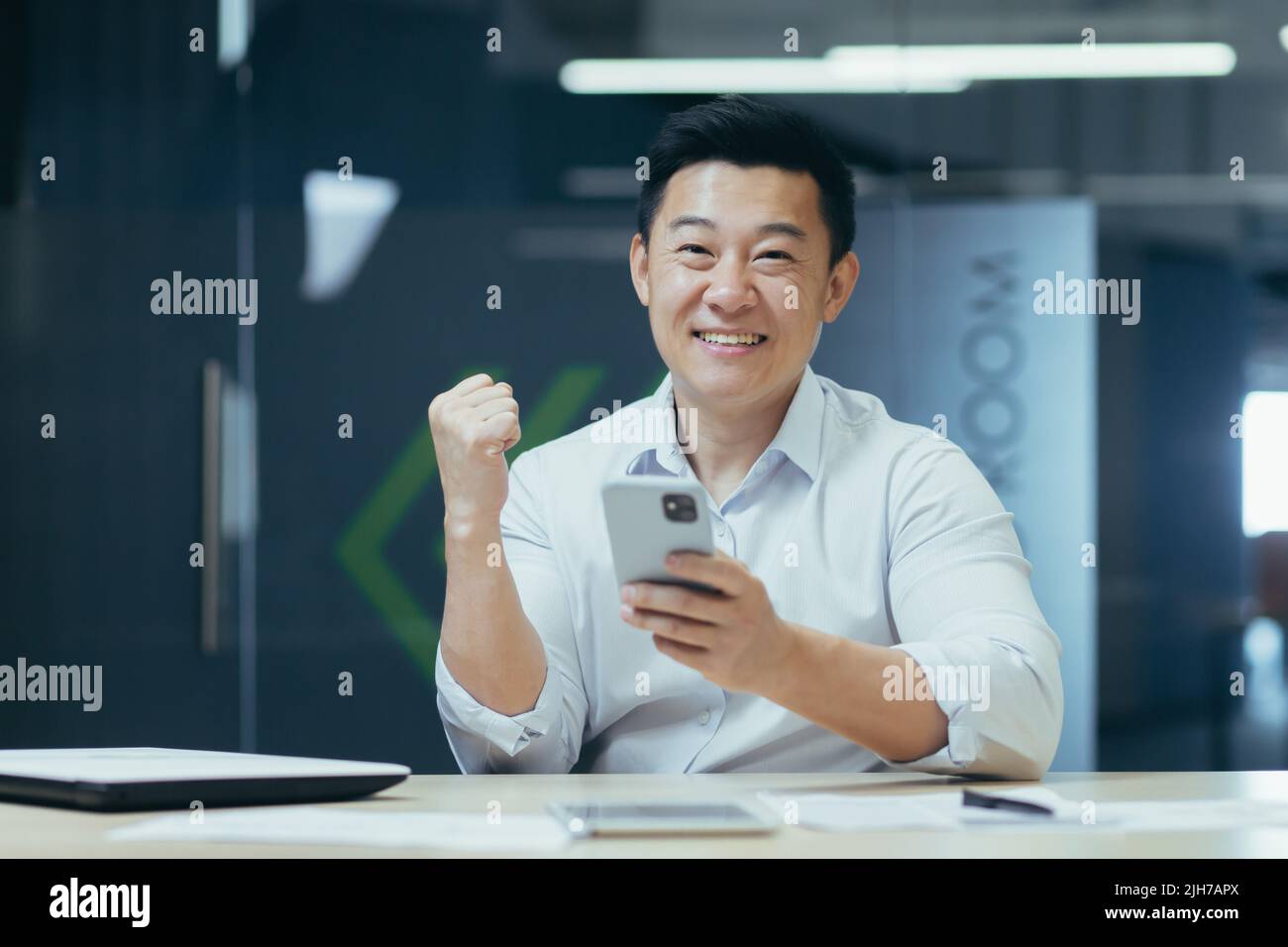 Successo vincente. Il giovane uomo d'affari asiatico tiene il telefono in mano, gioisce vincere, buone notizie, legge il messaggio. Sorridente. Si siede in un ufficio moderno a un tavolo con un notebook e un tablet, guardando la fotocamera Foto Stock