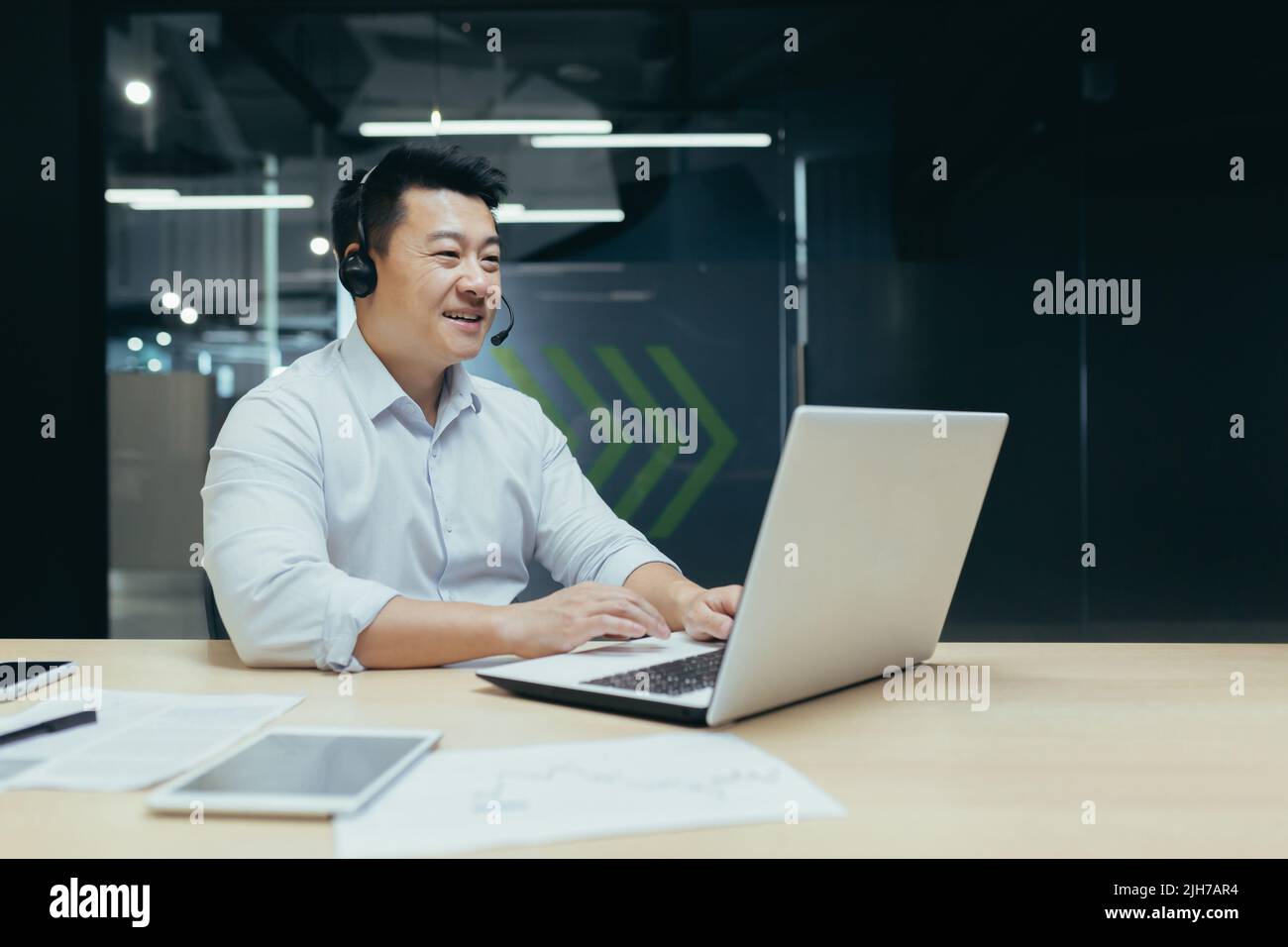 Un giovane uomo d'affari asiatico in cuffie con microfono si siede a un portatile in ufficio, conduce una conferenza online, un incontro remoto con i clienti, partner. Dice, spiega, sorride Foto Stock