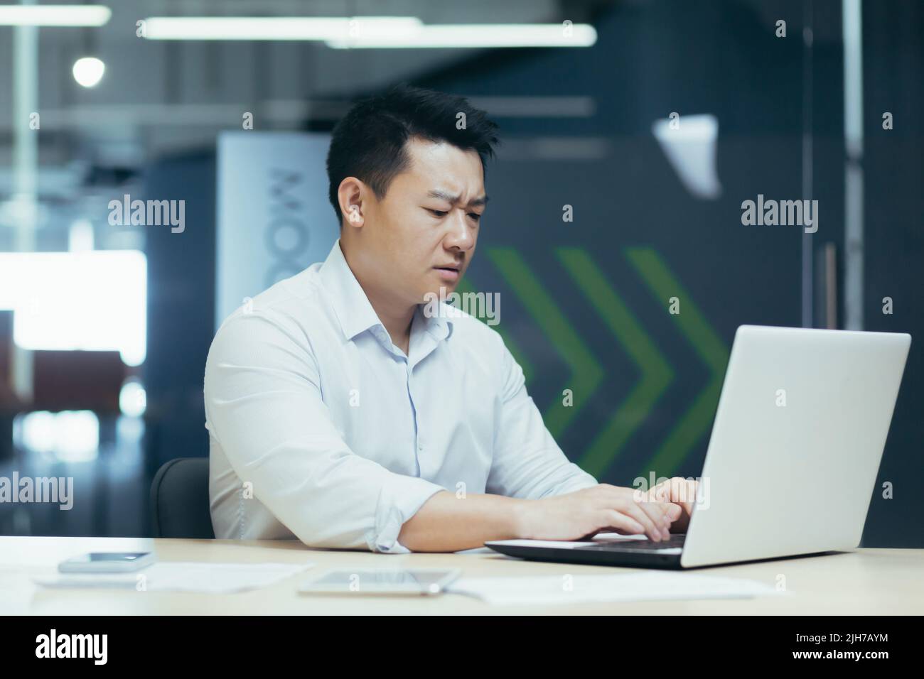 Serio e concentrato giovane uomo d'affari asiatico, libero professionista, imprenditore. Sedersi a un tavolo in un ufficio moderno, lavorare con un computer portatile e documenti. Foto Stock