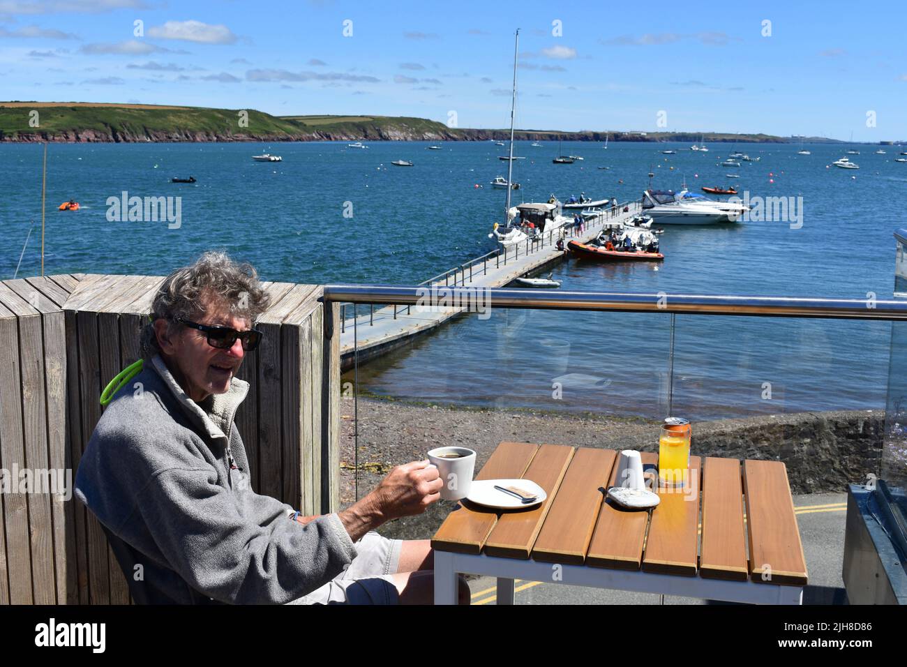 Uomo seduto con una tazza di caffè su una terrazza all'aperto con vista su Dale Bay, Dale, Pembrokeshire, Galles Foto Stock