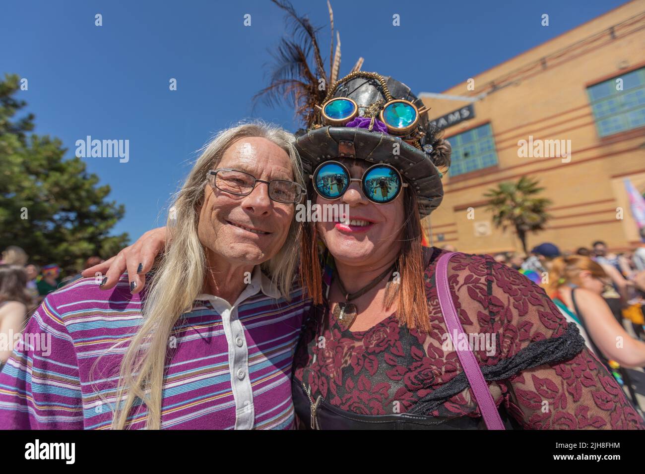 Southend on Sea, Regno Unito. 16 luglio 2022. Due persone posano insieme in un vivace evento all'aperto a Brighton. Uno indossa una polo a righe viola, mentre l'altro sfoggia un cappello piuma con occhiali protettivi, occhiali da sole riflettenti e una maglia con motivi neri e rossi. 1000 partecipanti si riuniscono per l'evento Southend Pride, prima che la parata inizi lungo High Street e nei Warrior Square Gardens per un Pride Festival con spettacoli dal vivo. Penelope Barritt/Alamy Live News Foto Stock