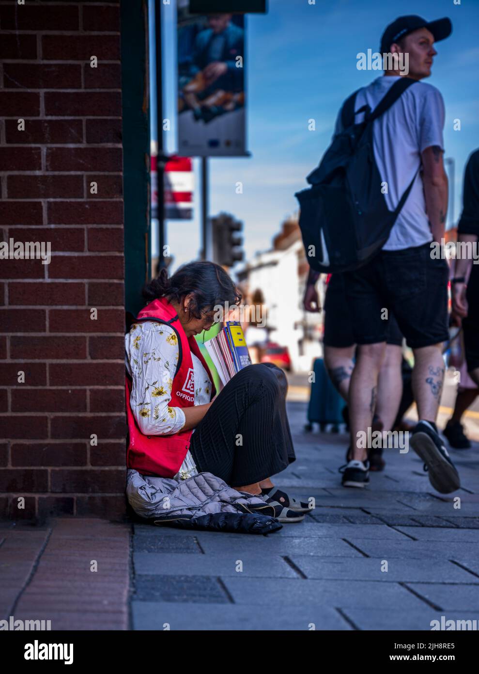 Grantham, Lincolnshire, Regno Unito – Una giovane donna asiatica sedeva sul marciapiede della strada vendendo copie di The Big Issue mentre la gente passava davanti e la ignorava Foto Stock