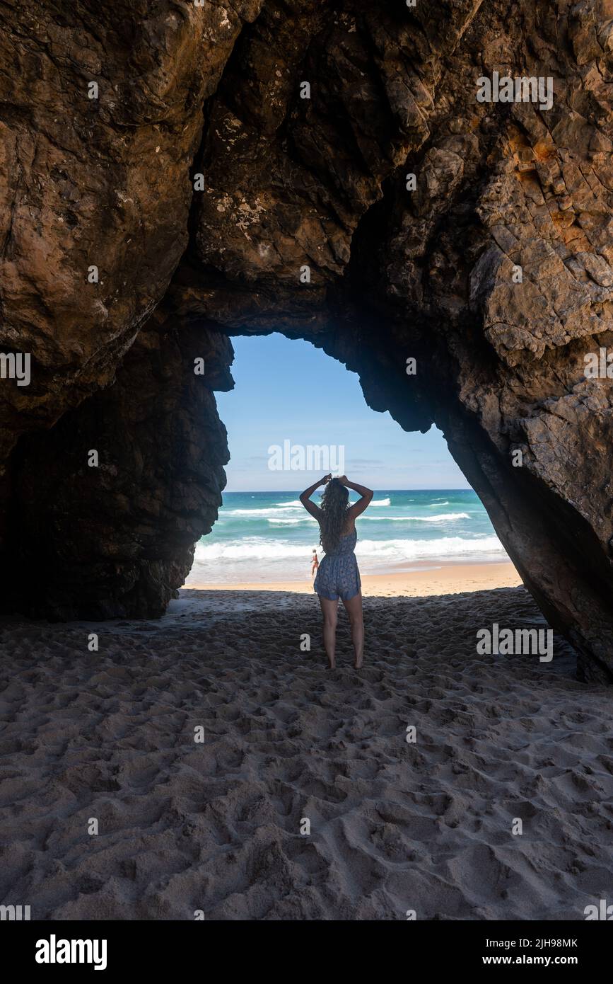 Silhouette della persona in un arco roccioso di grotta marina con spiaggia sabbiosa e oceano Atlantico oltre, costa di Sintra, Portogallo Foto Stock