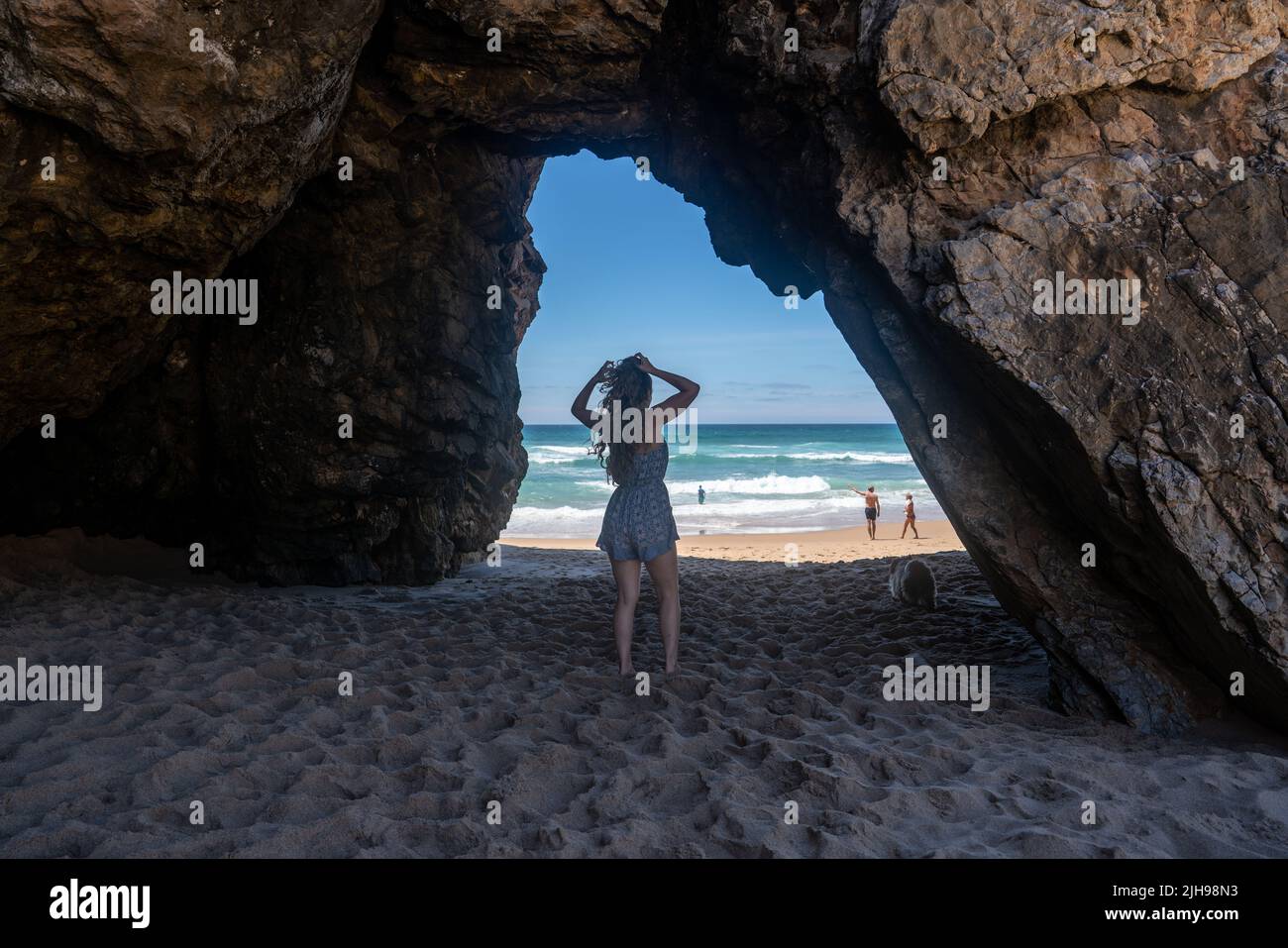 Donna in piedi in un suggestivo arco di grotta marina che incornicia la spiaggia sabbiosa e l'oceano Atlantico turchese, la costa di Sintra, Portogallo Foto Stock