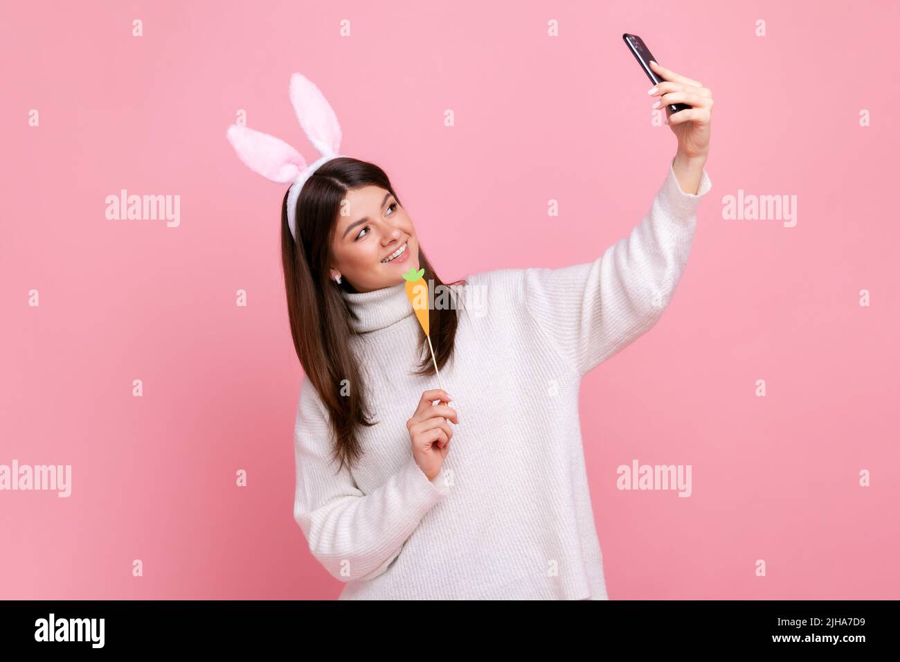 Ritratto di vista laterale di ragazza divertente con le orecchie conigliate in streaming o ha videochiamata con carota in mani, indossando bianco stile casual maglione. Studio interno girato isolato su sfondo rosa. Foto Stock