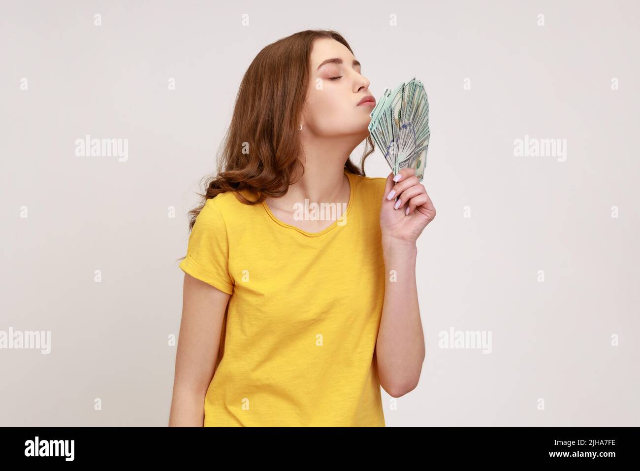 Ritratto della vista laterale di allegro ricco attraente giovane donna in giallo casual stile T-shirt in piedi e aria baciare fan di dollari, felice di vincere la lotteria. Studio interno girato isolato su sfondo grigio. Foto Stock