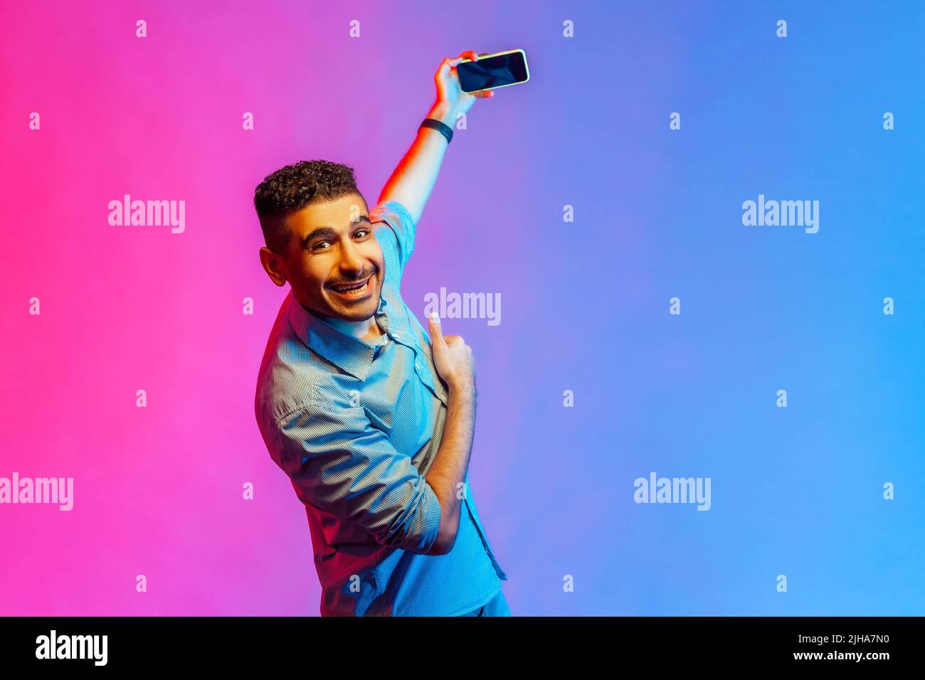 Ritratto di uomo felice sorridente in camicia in piedi con smartphone nelle mani, trasmissione in live streaming, guardare la fotocamera. Studio interno girato isolato su colorato sfondo di luce al neon. Foto Stock